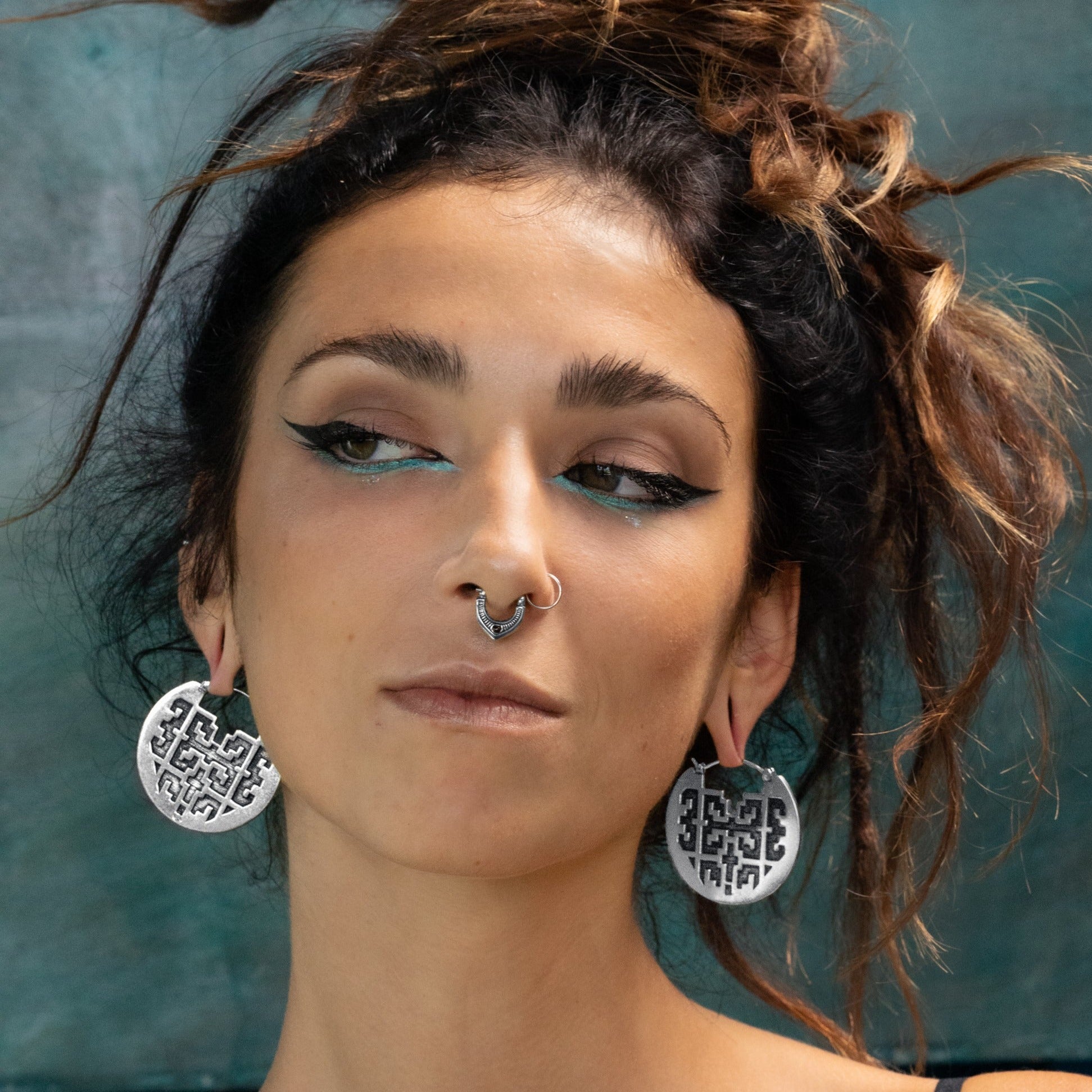 A woman with dark, curly hair is wearing large, round, silver CONIBOS Tribal Shipibo Hoop Disc Earrings in Silver by Kaya Movement and a nose ring. She has a winged eyeliner look, with turquoise accents on her lower eyelids. She is looking slightly to the side with a neutral expression against a textured green background.