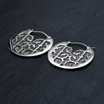 Introducing the CONIBOS Tribal Shipibo Hoop Disc Earrings in Silver by Kaya Movement: A pair of 16 gauge, round silver brass earrings with an intricate geometric cut-out design. Ideal for dark boho body jewelry enthusiasts, these tribal earrings are displayed on a dark, textured surface.