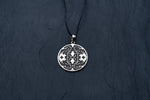 A round, intricately designed ICARO - White Brass Handmade Shipibo Icaros Pendant by Kaya Movement on a black cord lays on a dark, textured surface. The pendant features geometric patterns with oxidized details and a mix of black and silver tones, creating a maze-like appearance. The contrast highlights the design's intricate craftsmanship.