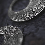 Close-up image of intricately designed OLIVIA Tribal Shipibo Seamless Disc Hoop Earrings in Silver by Kaya Movement, placed on a textured dark surface. The focus is on the detailed patterns etched into these 12 gauge earrings, which feature complex geometric and tribal motifs, adding a sense of elegance to the jewelry.