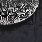 A close-up of the OLIVIA Tribal Shipibo Seamless Disc Hoop Earrings in Silver, designed by Kaya Movement, shows intricate raised geometric patterns on a dark, textured surface. The sharp lines and angles of the complex design are reminiscent of tribal art. The dark background contrasts sharply with the metallic surface of these 12-gauge earrings.