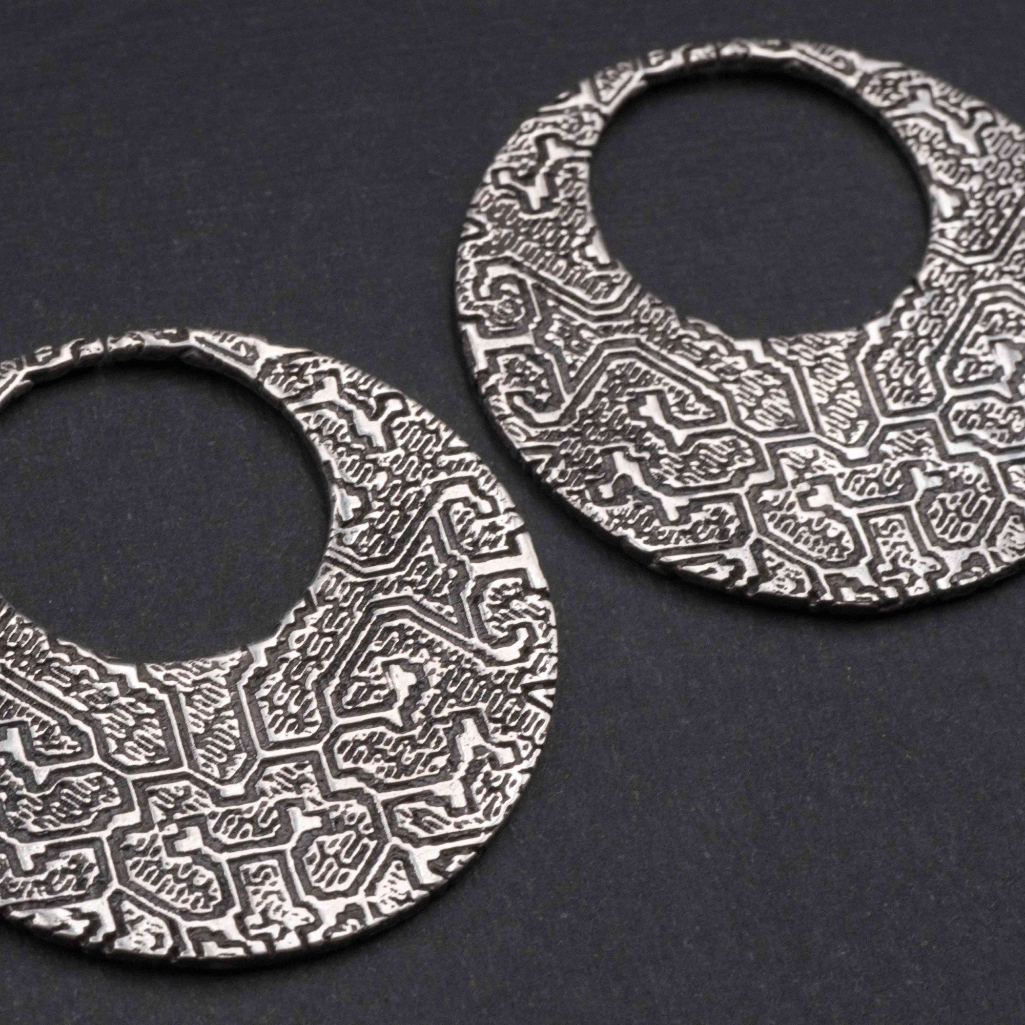 A pair of intricately designed OLIVIA Tribal Shipibo Seamless Disc Hoop Earrings in Silver from Kaya Movement lies on a dark surface. These 12 gauge earrings feature an ornate, geometric pattern etched throughout their surface with a circular cutout in the center, giving them a distinctive hoop-like appearance akin to boho statement earrings.