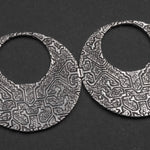 A close-up photo of two interconnected OLIVIA Tribal Shipibo Seamless Disc Hoop Earrings in Silver from Kaya Movement, featuring intricate geometric and tribal-like patterns. The black background highlights the detailed textures and designs on these 12-gauge boho statement earrings.