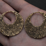 A person holds a pair of OLIVIA Tribal Shipibo Seamless Disc Hoop Earrings in Gold by Kaya Movement, which are crafted from brass and feature intricate black etchings. The 12 gauge earrings have a large circular cutout in the center and are placed on the person's palm for size reference. The dark background beautifully accentuates their tribal body jewelry design.