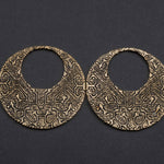 Introducing the OLIVIA Tribal Shipibo Seamless Disc Hoop Earrings in Gold by Kaya Movement. These 12 gauge gold brass earrings feature detailed geometric patterns and a large central cut-out, accentuating their hoop-like appearance. The dark background enhances their ornate design, capturing the essence of tribal body jewelry.