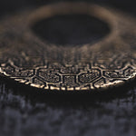Close-up of the OLIVIA Tribal Shipibo Seamless Disc Hoop Earrings in Gold by Kaya Movement, intricately detailed with geometric and patterned carvings, reminiscent of tribal body jewelry. The round metallic earrings lie on a dark surface, with the foreground in sharp focus and the background slightly blurred, emphasizing their texture and craftsmanship.