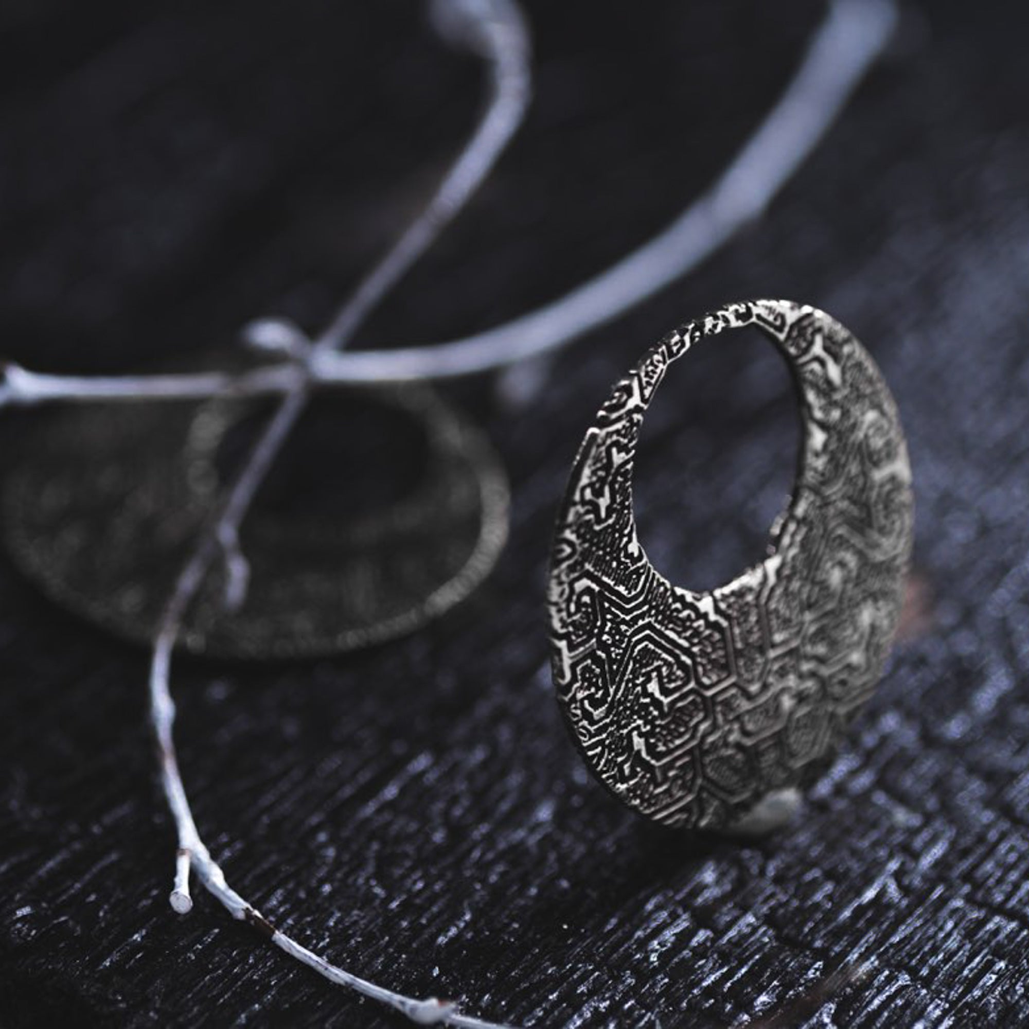 A close-up of an intricately designed OLIVIA Tribal Shipibo Seamless Disc Hoop Earring by Kaya Movement, featuring detailed patterns in 12 gauge silver, placed on a dark textured surface alongside a decorative branch. The earring's artistic craftsmanship truly stands out against the dark background.