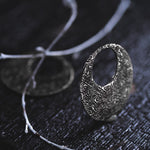 A close-up of an intricately designed OLIVIA Tribal Shipibo Seamless Disc Hoop Earring by Kaya Movement, featuring detailed patterns in 12 gauge silver, placed on a dark textured surface alongside a decorative branch. The earring's artistic craftsmanship truly stands out against the dark background.
