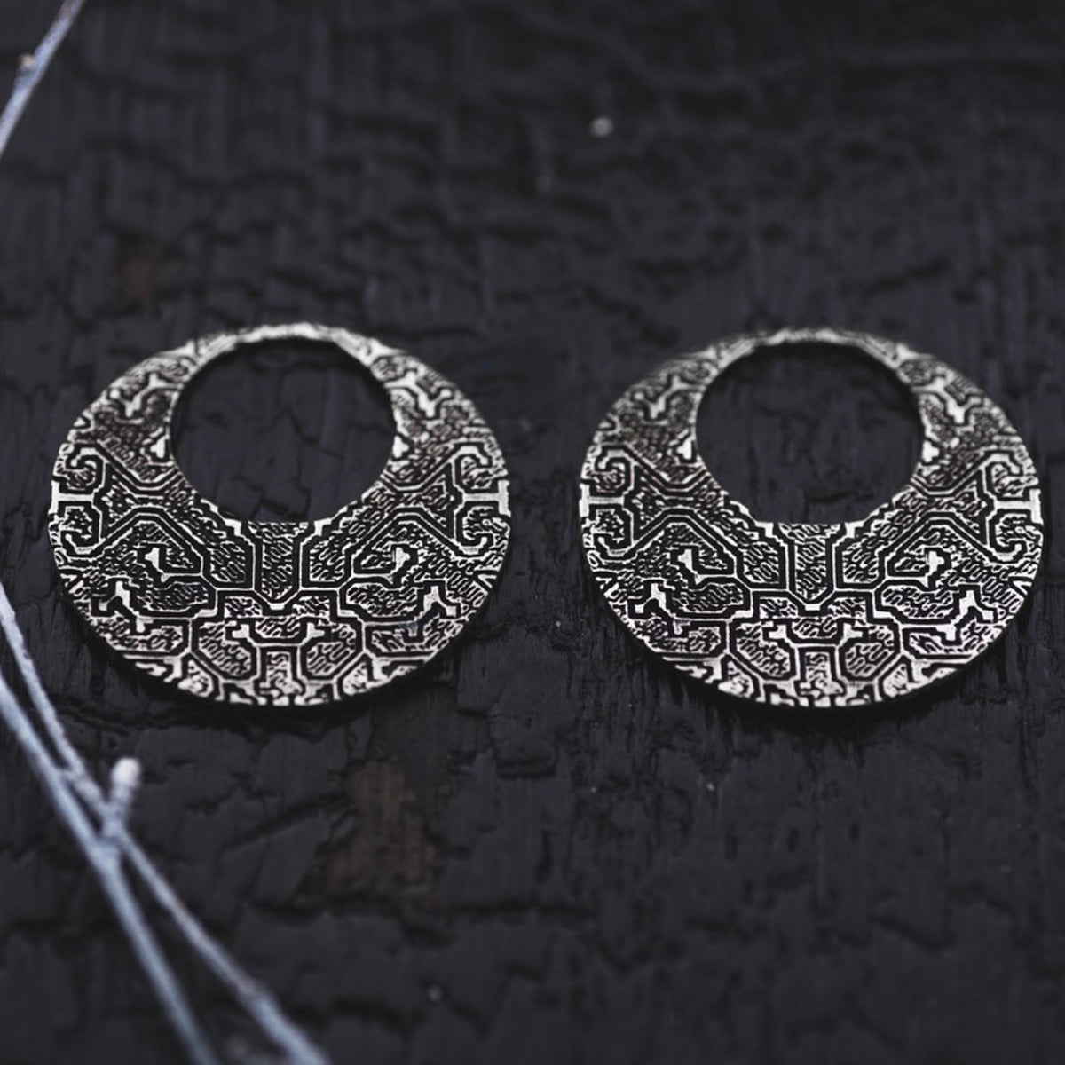 Placed on a dark textured surface, the OLIVIA Tribal Shipibo Seamless Disc Hoop Earrings in Silver from Kaya Movement exude a striking and artistic appearance with their detailed and symmetrical abstract design. These 12 gauge boho statement earrings showcase intricate engraving, while thin strands of white material are visible on the left side.