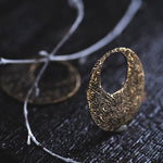 A close-up of an intricately designed OLIVIA Tribal Shipibo Seamless Disc Hoop Earring in gold by Kaya Movement, featuring a crescent shape and detailed patterns, placed on a dark textured surface. A blurred, similar piece of tribal body jewelry is visible in the background, partially obscured by crossing thin gray branches in the foreground.