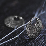 Close-up image of Kaya Movement's KONI Tribal Shipibo Endless Hoop Disc Earrings in Silver, showcasing an 18-gauge intricately patterned circular earring, possibly tribal body jewelry, resting on a dark textured surface with branches in the background. Made from 925 sterling silver, the earring features elaborate maze-like designs that add an artistic and detailed appearance.