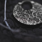 A close-up image of the KONI Tribal Shipibo Endless Hoop Disc Earrings in Silver by Kaya Movement resting on a dark, textured surface. These earrings, designed as tribal body jewelry, feature detailed abstract patterns and have a circular shape with a hole at the top for attaching.