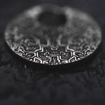 Close-up image of a round, intricately engraved metal object, possibly the KONI Tribal Shipibo Endless Hoop Disc Earrings in Silver by Kaya Movement, laying on a textured, dark surface. The detailed engravings depict geometric patterns and mythical elements reminiscent of tribal body jewelry. There is a small hole near the top of the object.