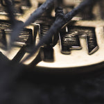 Close-up image of the CONIBOS Tribal Shipibo Hoop Disc Earrings in Gold by Kaya Movement, partially obscured by dark, blurred branches or wires in the foreground. The earrings feature intricate engravings reminiscent of tribal designs that are partially visible. The background is dark and out of focus, enhancing the earrings' texture and shine.
