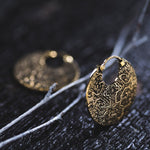 Close-up of the Kaya Movement KONI Tribal Shipibo Endless Hoop Disc Earring in Gold, intricately designed with geometric patterns, secured around a thin branch. A second, similar tribal earring is blurred in the background on a dark, textured surface.