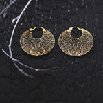 A pair of KONI Tribal Shipibo Endless Hoop Disc Earrings in Gold by Kaya Movement rests on a black textured surface. Each earring features intricate, symmetrical etchings. A light-colored branch lies across the left side of the image, contrasting with the dark background.
