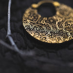 Close-up of Kaya Movement's KONI Tribal Shipibo Endless Hoop Disc Earring in gold, intricately patterned and resting on a textured black surface. This 18 gauge earring features detailed designs, partially in focus to highlight its ornate craftsmanship. The background includes a blurred white twig.