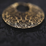 Close-up of a round, intricately patterned gold earring with a hole at the top. The surface of the KONI Tribal Shipibo Endless Hoop Disc Earring in Gold by Kaya Movement features detailed geometric and possibly symbolic designs reminiscent of boho statement pieces. The background is dark and slightly textured, providing contrast to the golden hues of the earring.