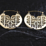 Close-up view of the Kaya Movement CONIBOS Tribal Shipibo Hoop Disc Earrings in Gold, featuring intricate geometric cut-out designs, placed on a black textured surface. The earrings have symmetrical patterns and thin wires attached to the top for wearing. These earrings are 16 gauge in size.