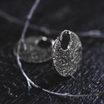 A close-up of a KONI Tribal Shipibo Endless Hoop Disc Earring in Silver (18 gauge) from Kaya Movement, showcasing an intricately designed, textured circular pattern resting on a dark wooden surface. A delicate twig lies across the earring, enhancing the artistic and natural feel of the composition, perfectly evoking Kaya Movement's essence. The background is out of focus.
