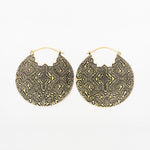 A pair of Kaya Movement's oversized PANCHA Tribal Shipibo Disc Earrings in gold, designed with intricate geometric patterns etched on the surface. These large round metallic earrings feature interlocking shapes and lines, creating a complex, ornamental appearance. The earrings have hoop clasps at the top and are made with 16 gauge material.