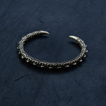 A close-up of the SHIRO Men's Gold Cuff Bracelet by Kaya Movement, showcasing its intricately designed dark and textured pattern resembling a reptile spine against a black slate background. This adjustable gold cuff features two sharp, golden tips on either end and is adorned with striking geometric shapes.