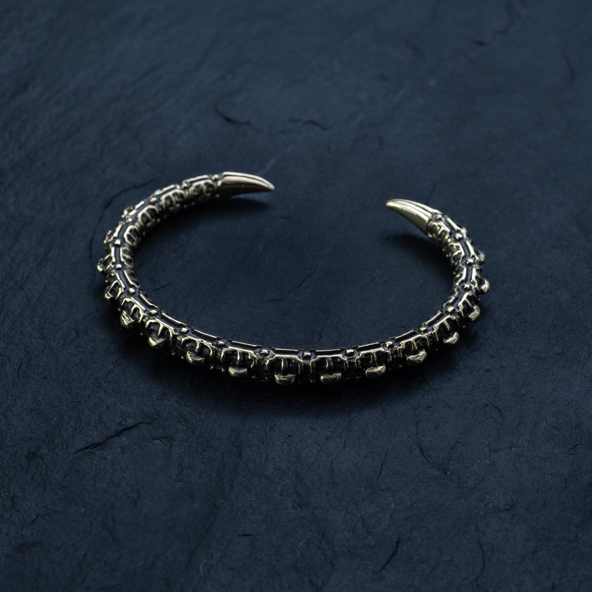 A close-up of the SHIRO Men's Gold Cuff Bracelet by Kaya Movement, showcasing its intricately designed dark and textured pattern resembling a reptile spine against a black slate background. This adjustable gold cuff features two sharp, golden tips on either end and is adorned with striking geometric shapes.