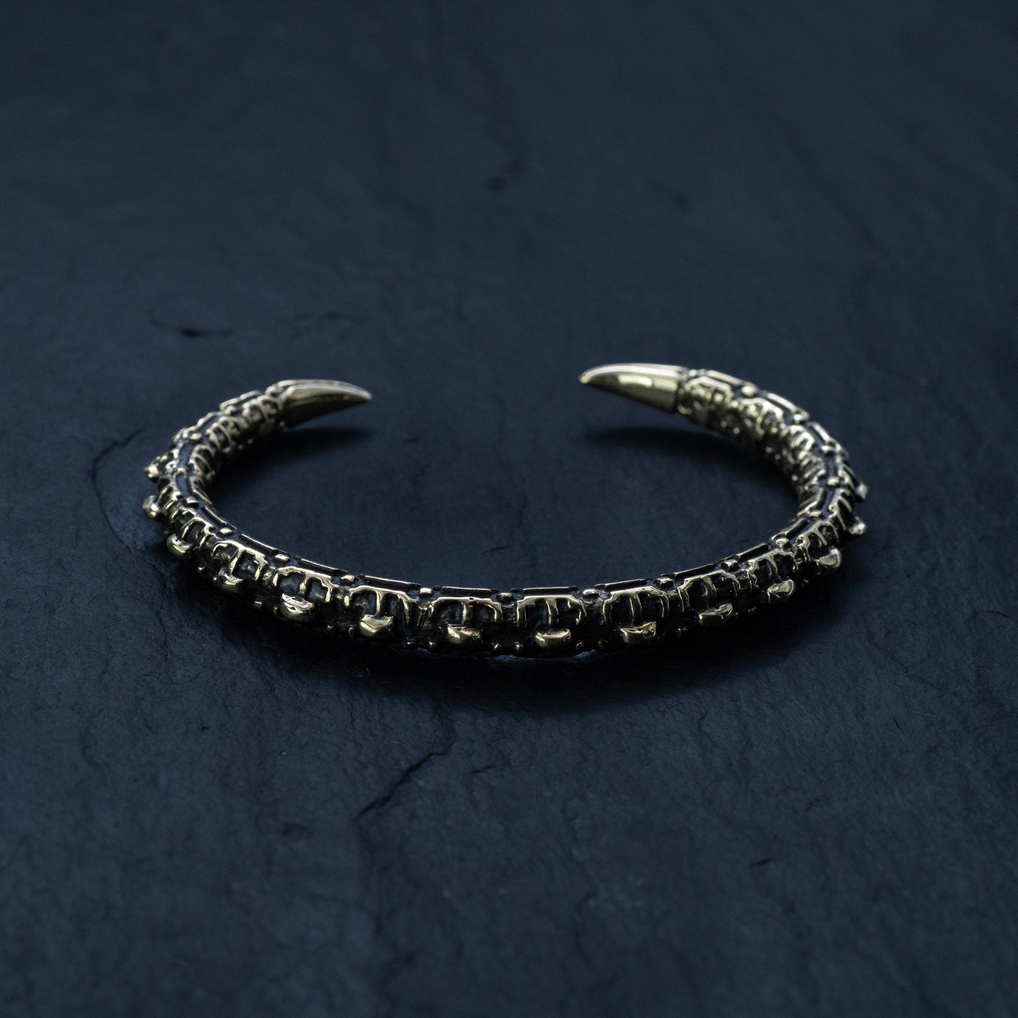 A close-up image of the SHIRO Men's Gold Cuff Bracelet by Kaya Movement, photographed on a dark slate surface. The intricately designed adjustable cuff features a segmented, textured pattern resembling a reptile's spine and claw-like ends that point towards each other, forming a semi-circle.
