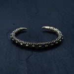 A close-up image of the SHIRO Men's Gold Cuff Bracelet by Kaya Movement, photographed on a dark slate surface. The intricately designed adjustable cuff features a segmented, textured pattern resembling a reptile's spine and claw-like ends that point towards each other, forming a semi-circle.