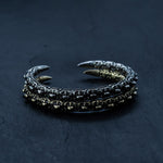 Two intricately designed SHIRO Bangles from Kaya Movement resemble claws. One is crafted from dark metal, possibly oxidized silver, and the other boasts a golden hue. Both men's cuff bracelets feature chain-like segments along their length with sharp claw-like ends, all placed on a dark surface.