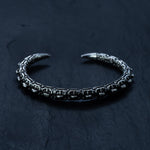 The SHIRO Bangle in Silver by Kaya Movement, an intricately designed men's cuff bracelet with a slightly open end, is laid on a dark, textured surface. The oxidized silver bracelet features a pattern resembling interlocking Shipibo links, and the ends taper into smooth, rounded points.
