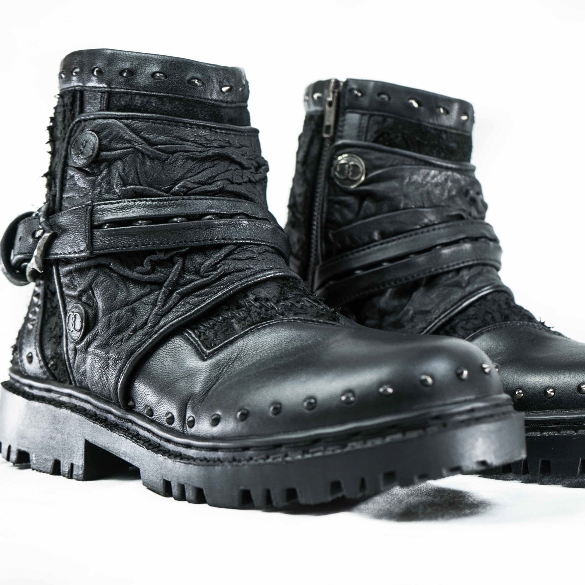 Presenting the MAGMA Wasteland Studded Leather Ankle Combat Boots by Kaya Movement: a pair of black, rugged, leather lace-up punk rock boots featuring textured details, metal studs, straps, and a side zipper. These boots boast intricate stitching and a thick, durable sole designed for heavy-duty wear. The backdrop is plain white.