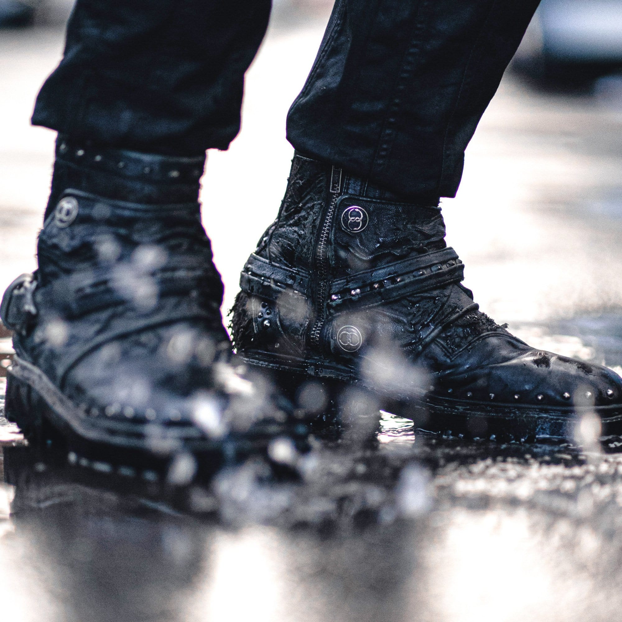 Close-up of a person wearing Kaya Movement's MAGMA Wasteland Studded Leather Ankle Combat Boots, featuring metal accents and textured details, standing on a wet surface with splashes of water around. The individual is dressed in black pants, suggesting rainy or wet conditions.