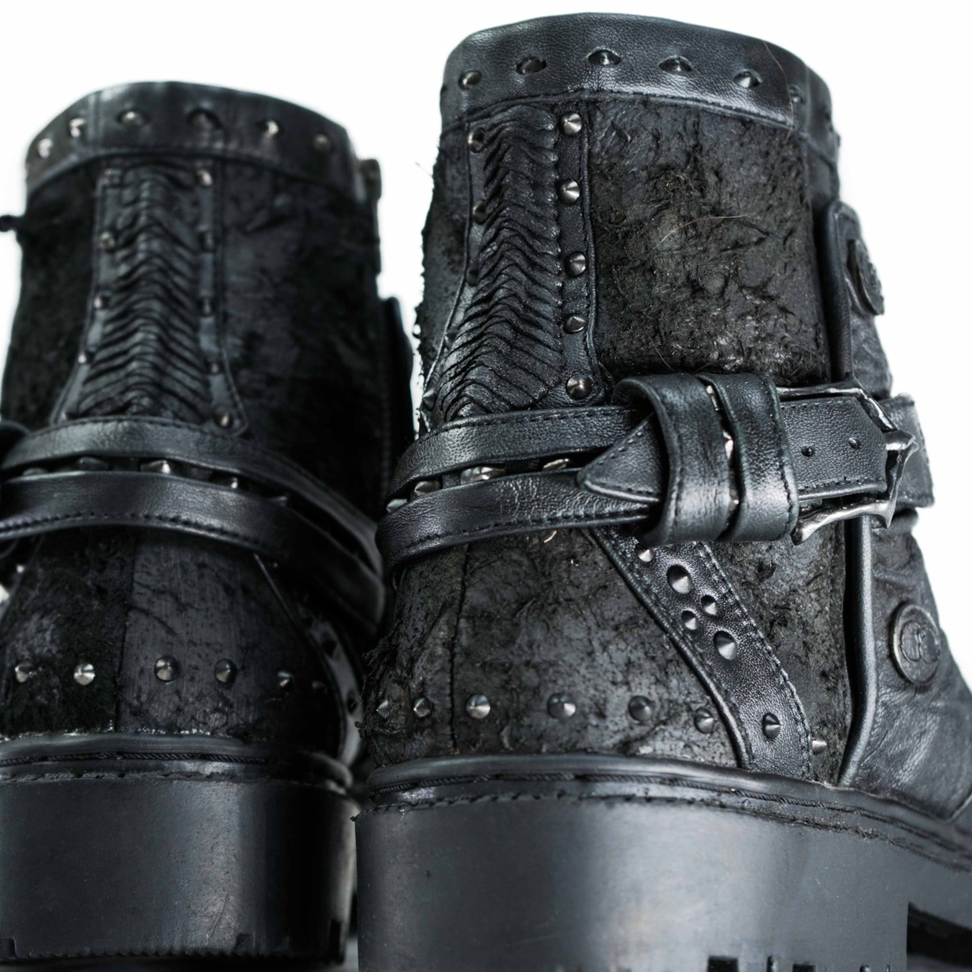 Close-up view of the MAGMA Wasteland Studded Leather Ankle Combat Boots by Kaya Movement. These black, textured boots showcase studded details along with decorative straps and buckles around the ankles, offering a rugged yet stylish design. The thick soles further enhance their robust appearance.