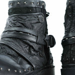 Close-up of a pair of worn, black leather boots with a rugged design. These MAGMA Wasteland Studded Leather Ankle Combat Boots from Kaya Movement feature decorative metal studs, textured leather, straps, and buckles. The material appears distressed, giving the studded leather ankle boots a unique, vintage look. The background is white.