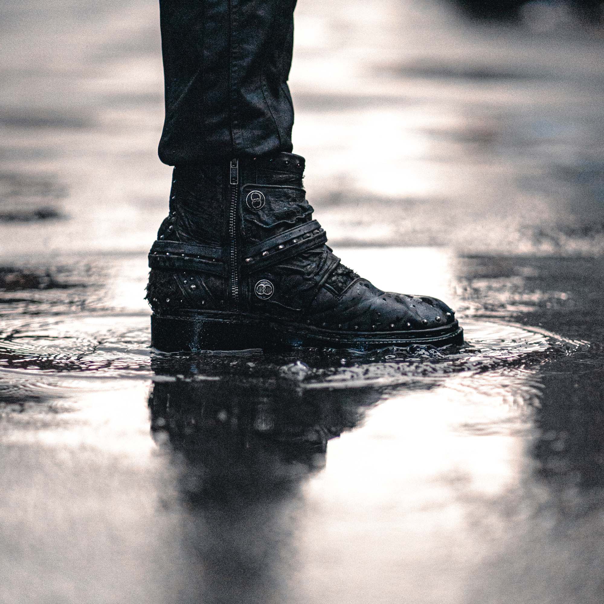 A person wearing black leather pants and punk rock boots stands in a puddle on a shiny wet street. The Kaya Movement MAGMA Wasteland Studded Leather Ankle Combat Boots feature zippers, multiple straps with metal accents, and reflect their rugged style in the water below.