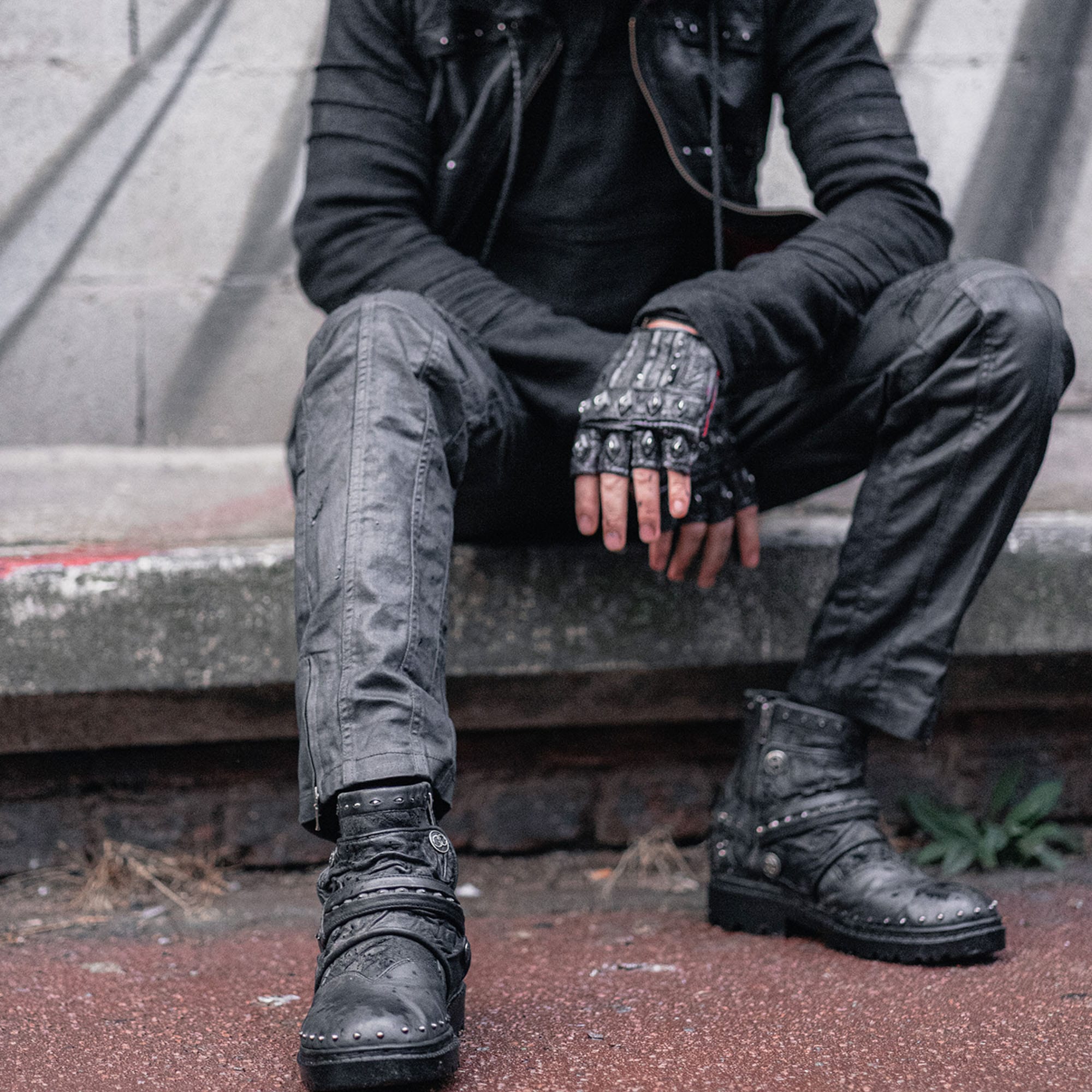 A person dressed in punk-inspired attire sits on a concrete ledge. They wear Kaya Movement's MAGMA Wasteland Studded Leather Ankle Combat Boots, dark grey pants, a black jacket with metal details, and fingerless gloves. The person's face is not visible, and the background includes a graffiti-covered wall.