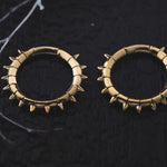 Close-up of the KALASH Medium Spike Hoop Clicker Ear Weights in Gold Brass by Kaya Movement, designed with small spikes along their outer edges, and placed on a dark textured surface. Thin white lines intersect the corners of the image, adding a delicate contrasting element. Perfect for those with stretched ear lobes at 4 gauge size.
