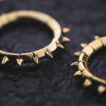 Two KALASH Medium Spike Hoop Clicker Ear Weights in Gold Brass by Kaya Movement are displayed on a textured dark surface. Each 4-gauge ear weight features evenly spaced small spikes around its circumference, giving them a bold, edgy appearance. The weights are positioned side by side, with one slightly overlapping the other.