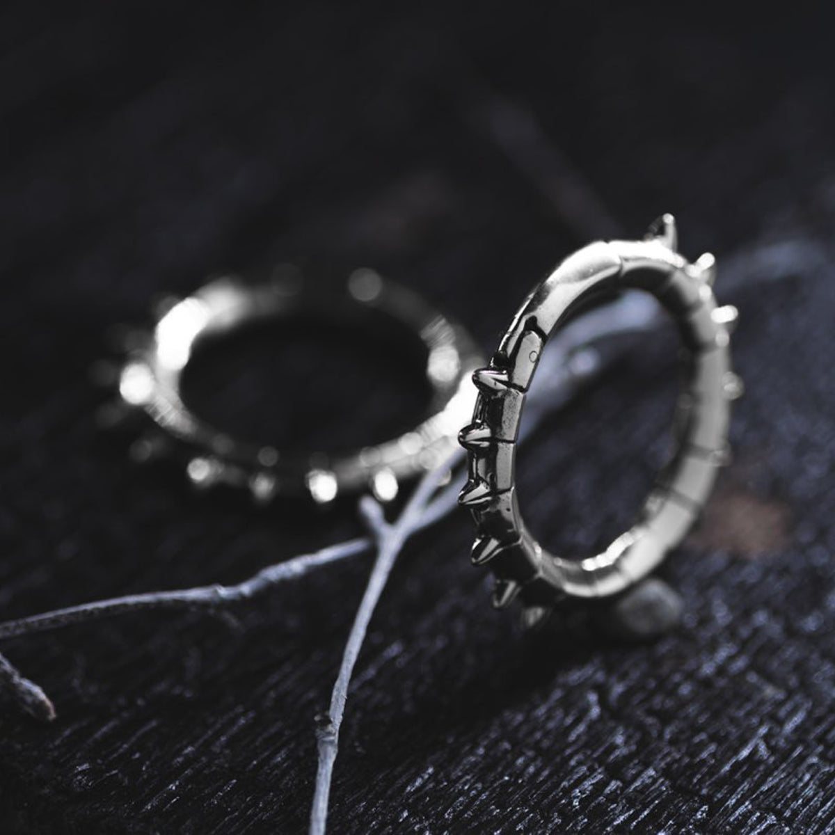 Dark-themed photograph featuring KALASH Medium Spike Hoop Clicker Ear Weights in Silver Brass by Kaya Movement, exuding a tribal punk industrial jewelry vibe. One ear weight lies flat on a dark surface while the other stands upright, accompanied by dry, thin twigs that add a contrasting natural element.