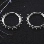 Close-up of a pair of Kaya Movement's KALASH Medium Spike Hoop Clicker Ear Weights in Silver Brass (4 gauge), adorned with small spikes around the edges, set against a textured black background. These tribal punk industrial jewelry pieces add a bold, edgy aesthetic to any outfit.