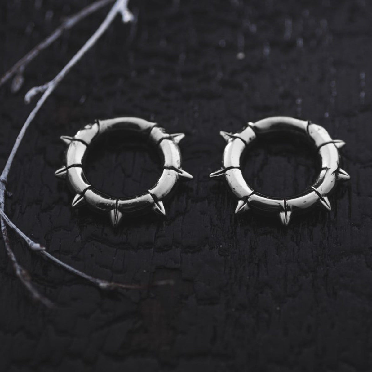 Two KALASH Small Spike Hoop Clicker Ear Weights in Silver Brass by Kaya Movement with evenly spaced spikes lie on a textured, dark surface. Thin, delicate twigs are visible on the left side of the image, adding a contrasting natural element to the scene.