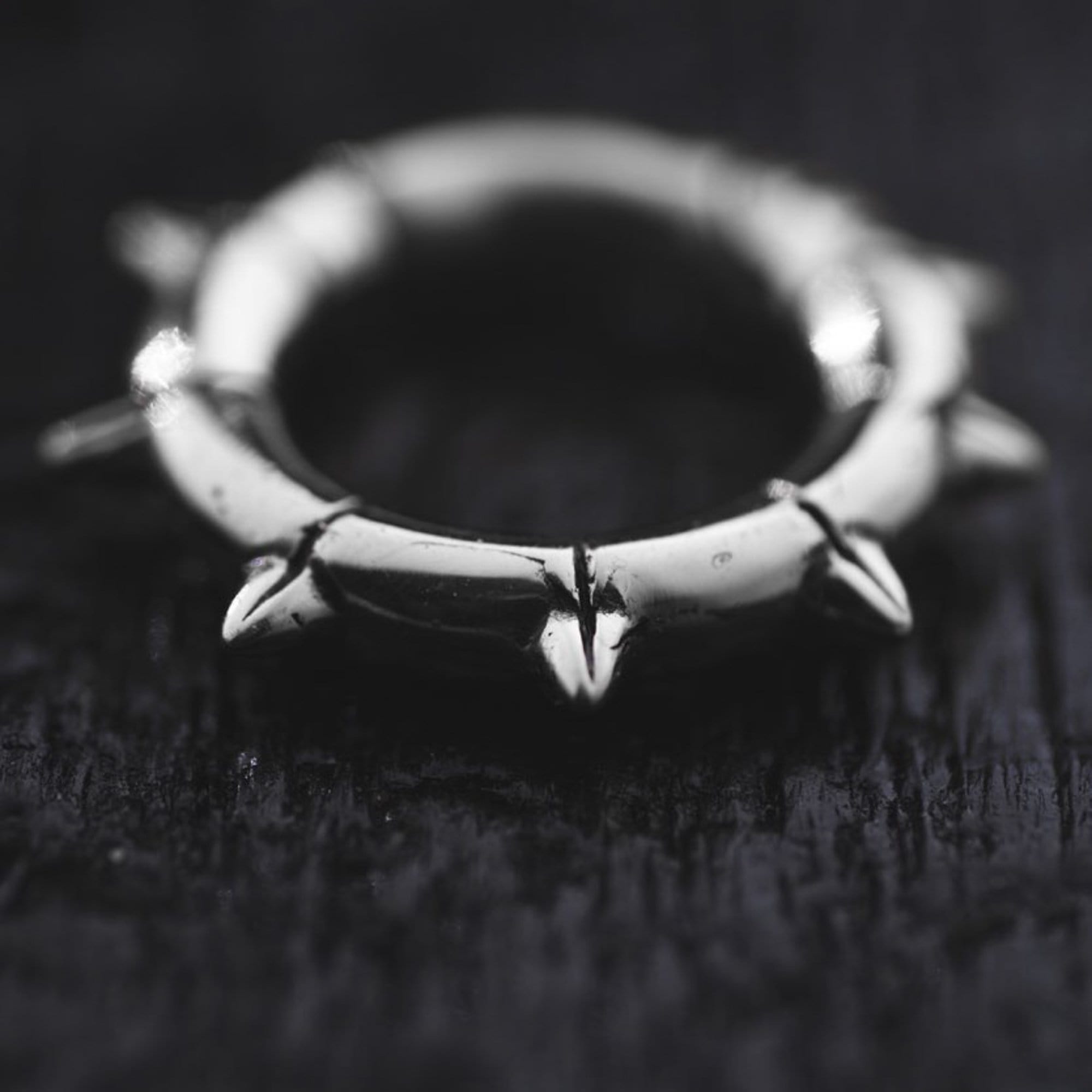 Close-up of a KALASH Small Spike Hoop Clicker Ear Weights in Silver Brass by Kaya Movement, featuring thorn-like protrusions evenly spaced around its outer edge, resembling a biomechanical hoop clicker, resting on a dark, textured surface. The piece could easily be mistaken for unisex 4 gauge earrings.