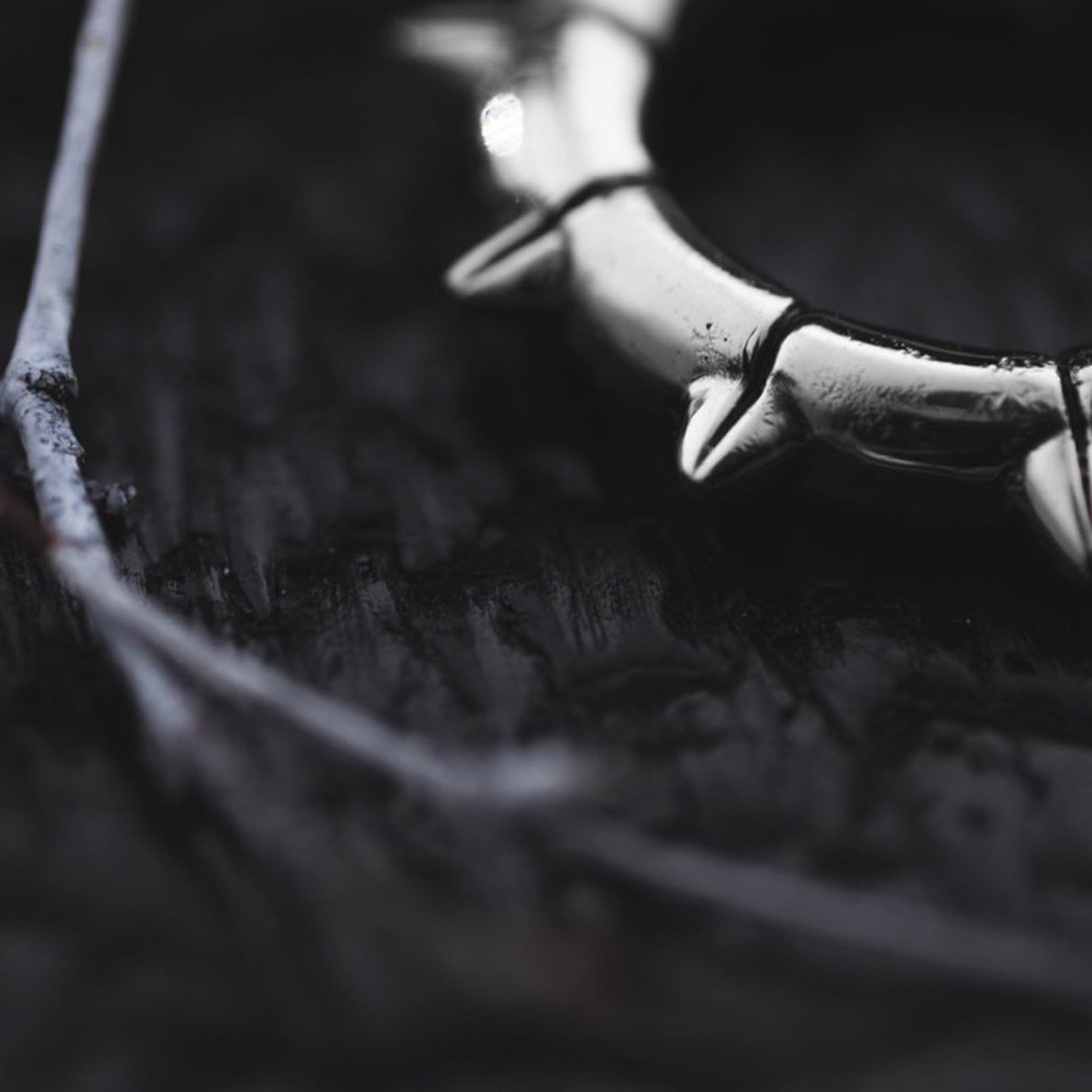 Close-up of the KALASH Small Spike Hoop Clicker Ear Weights in Silver Brass by Kaya Movement, resting on a dark, textured surface. A thin twig lies nearby, partially out of focus. The image captures a moody and dramatic atmosphere with its dark tones and selective focus, evoking the edgy allure often seen in unisex gauge earrings.
