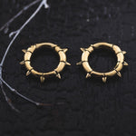 A close-up photo of two KALASH Small Spike Hoop Clicker Ear Weights in Gold Brass by Kaya Movement, designed with small spikes evenly spaced around their circumference. The ear weights, resembling post-apocalyptic body jewelry, are placed side by side on a dark, textured surface with a thin, white twig beside them.