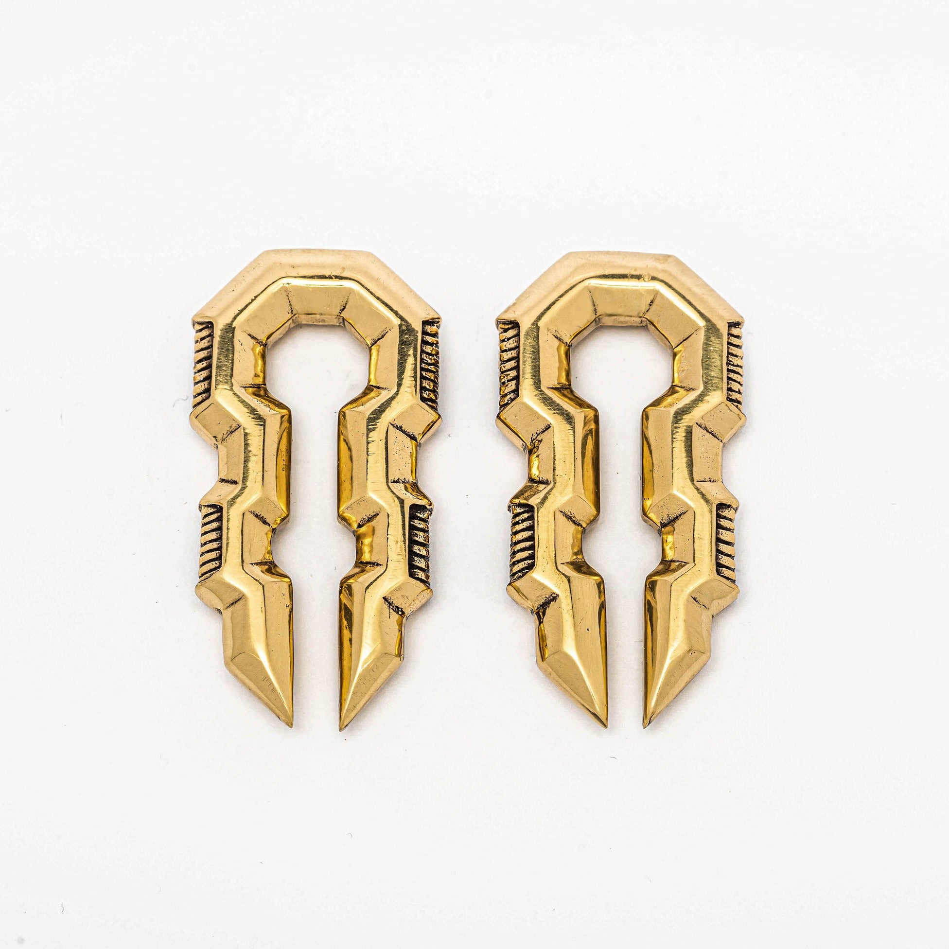 Introducing the TECHRE Futuristic Sci Fi Hexagon Keyhole Ear Weights in Gold by Kaya Movement. These bold, golden ear weights feature a sleek geometric design with sharp edges and angular shapes that create an elongated, symmetrical pattern, reminiscent of biomechanical cyborg hangers. The 0-gauge earrings are showcased against a plain white background.