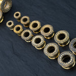 A collection of TERA Futuristic Sci Fi Flesh Tunnels in Gold Brass by Kaya Movement is artfully arranged in a triangular formation on a dark slate surface. The tunnels range from 8mm to 20mm, with the largest at one end and the smallest at the other, highlighting exquisite body jewelry craftsmanship.