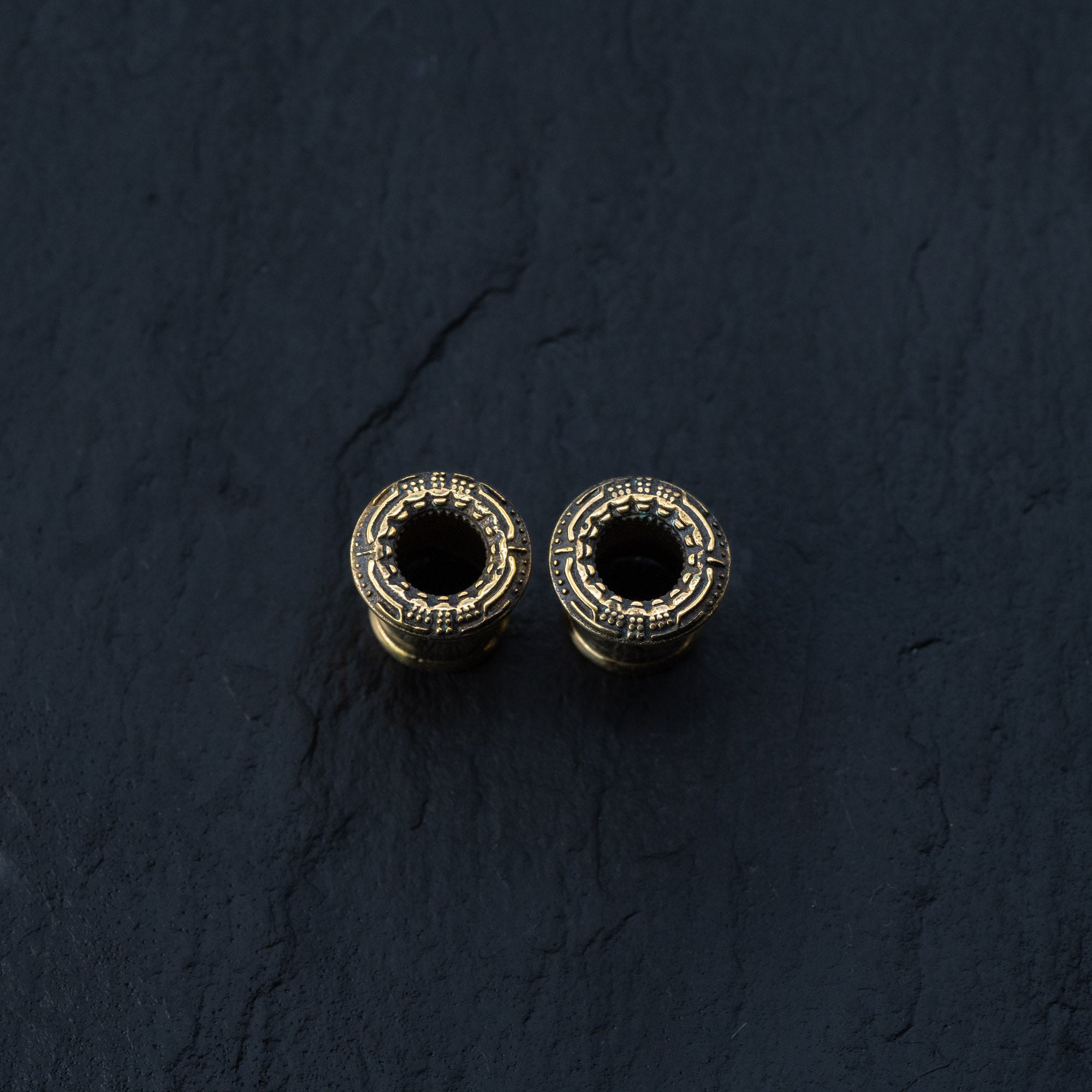 A striking pair of TERA Futuristic Sci Fi Flesh Tunnels in Gold Brass by Kaya Movement, featuring ornate, detailed patterns on a dark, textured surface. These hollow cylindrical objects are available in sizes ranging from 8mm to 20mm and are perfect for body jewelry enthusiasts who appreciate futuristic designs.