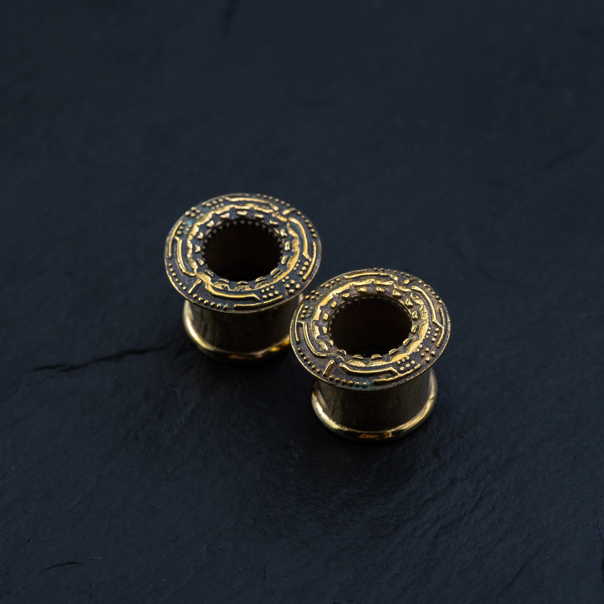 Close-up shot of two Kaya Movement's TERA Futuristic Sci Fi Flesh Tunnels in gold brass, featuring intricate patterns on their exposed circular tops. They are displayed on a black, textured surface, showcasing their hollow centers.