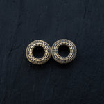 A close-up image of two TERA Futuristic Sci Fi Flesh Tunnels in gold brass by Kaya Movement, placed side by side on a dark, textured surface. These intricate pieces feature detailed patterns resembling small symbols or engravings encircling a hollow center and are available in sizes ranging from 8mm to 20mm.